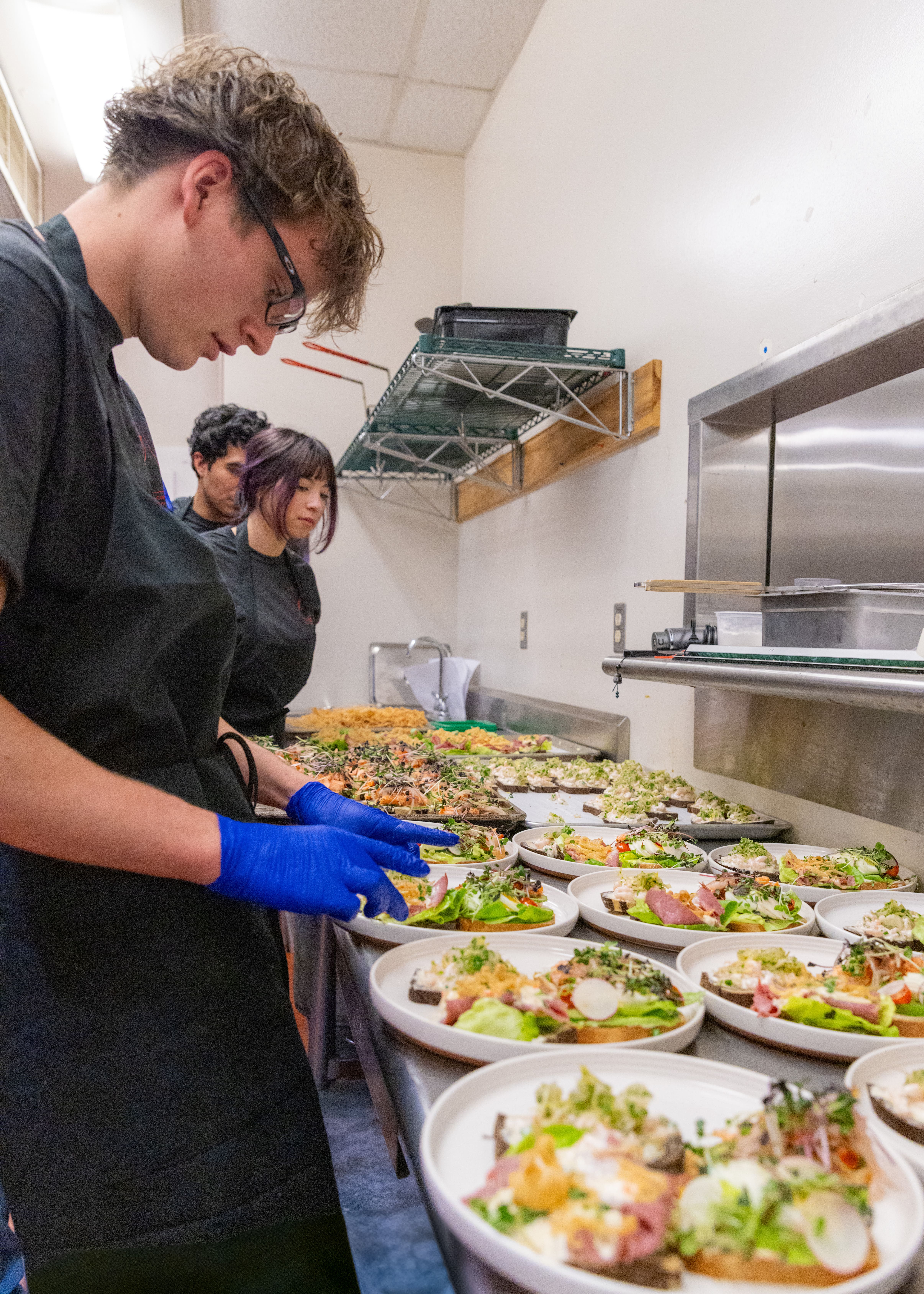 Young chefs in a kitchen prepare dishes with focus, wearing black aprons and gloves. Plates of colorful salads line the counter, creating a lively atmosphere.