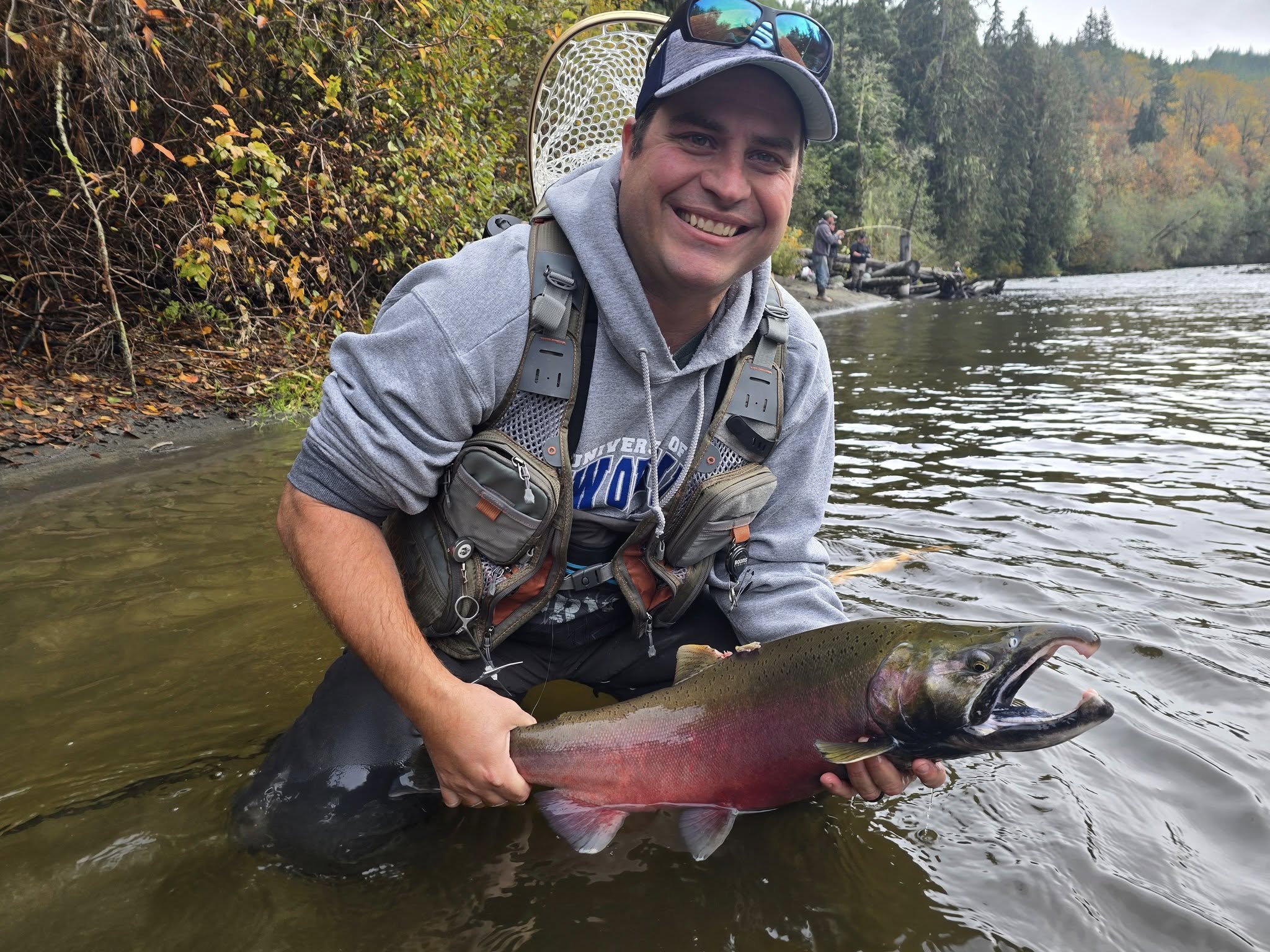 Jon McKenize smiling while holding a salmon in a river with trees in the background