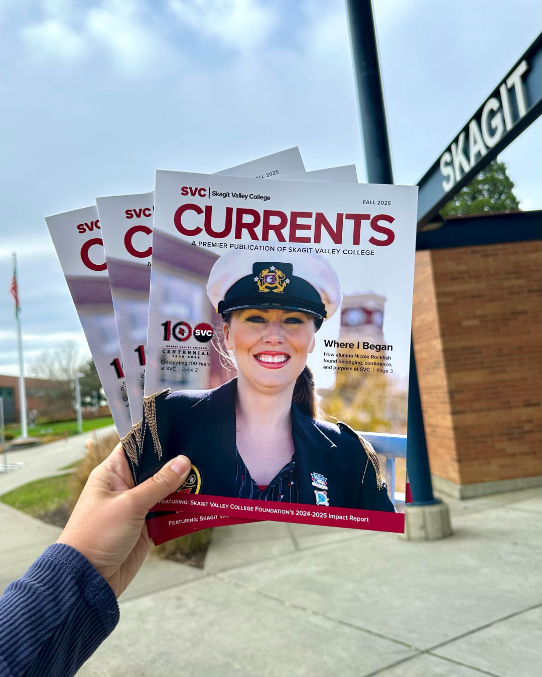 2025 Fall Currents Available Now A hand holding copies of "Currents," featuring a smiling woman in a naval uniform on the cover. The Skagit Valley College sign is visible in the background.